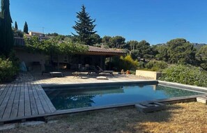 Une piscine avec une terrasse en bois, un patio couvert et une vue sur une colline arborée.