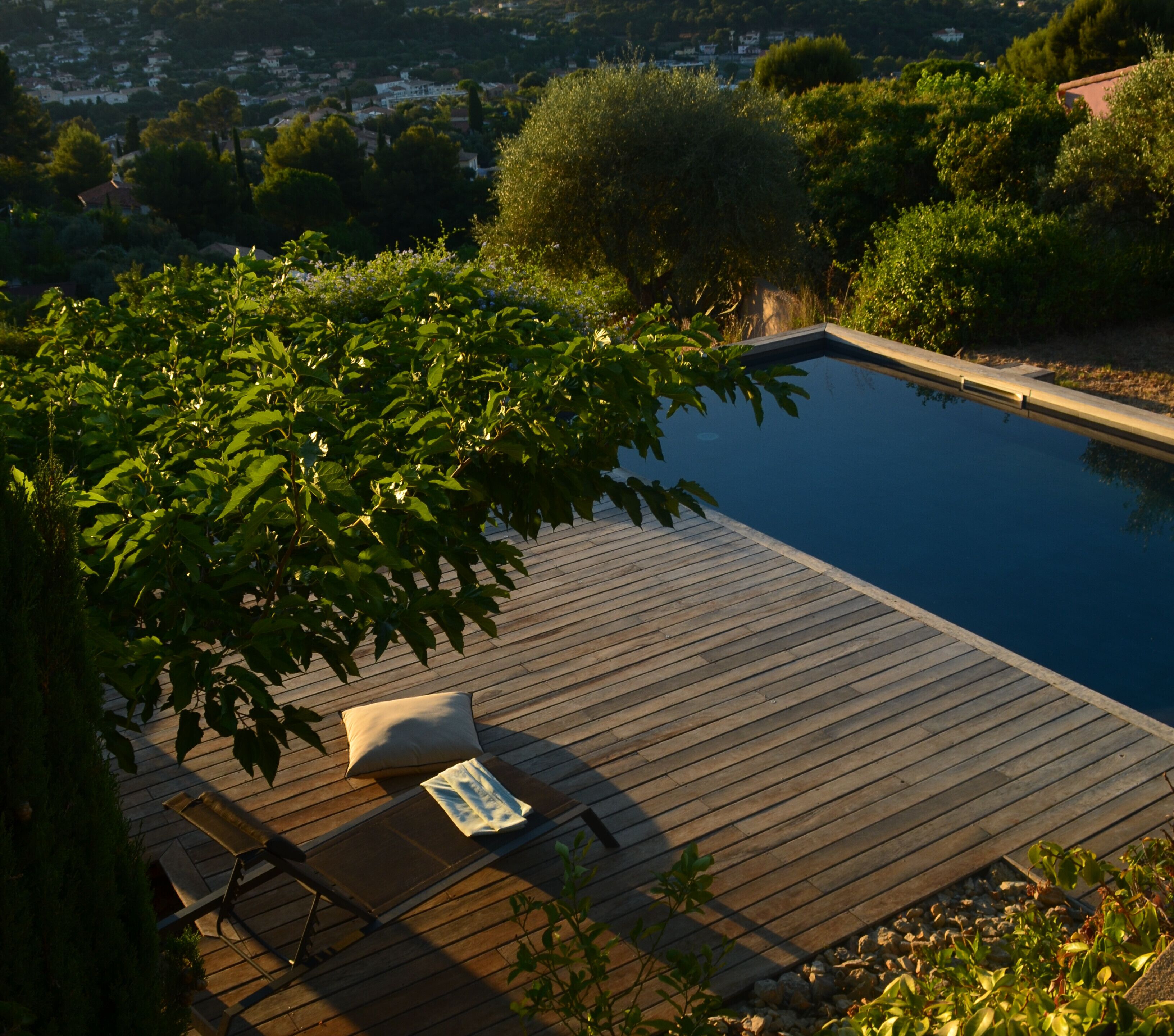A wooden deck with a pool, surrounded by greenery and overlooking a residential area.