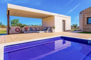 Un espace piscine extérieur moderne, doté d’une terrasse en bois, d’un coin salon intégré avec des coussins et, en arrière-plan, d’un bâtiment en pierre.