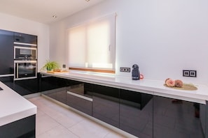 A modern kitchen with black cabinets, a white countertop, a built-in oven, a coffee machine, and a window with blinds. - Villa Pareteta - Three Bedroom Villa, Sleeps 6 (Binissalem)