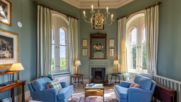 A room with two blue armchairs, a fireplace, a chandelier, and large windows with curtains.