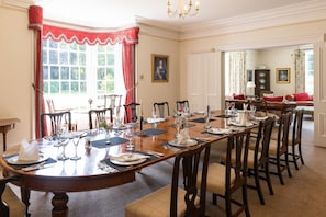 A dining room with a long wooden table set for a meal, flanked by chairs, with a view of a garden through the window.