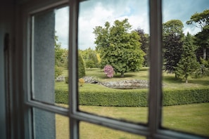 A view through a window of a well-maintained garden with a variety of trees, a stone bridge, and neatly trimmed hedges.