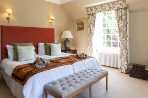 A hotel room with a large bed, a bench, a nightstand, a lamp, a window with curtains, and a radiator.