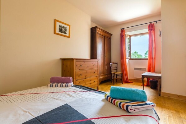 Quarto com cama, cómoda de madeira, cadeira, janela com cortinas e uma pequena mesa.