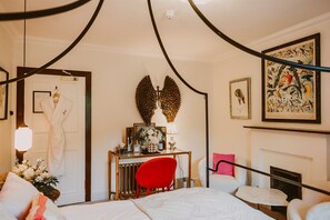 A hotel room with a bed, a desk with a lamp, a red chair, a fireplace, and framed artwork on the walls.