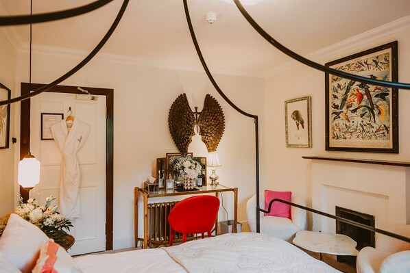 A hotel room with a bed, a desk with a lamp, a red chair, a fireplace, and framed artwork on the walls.