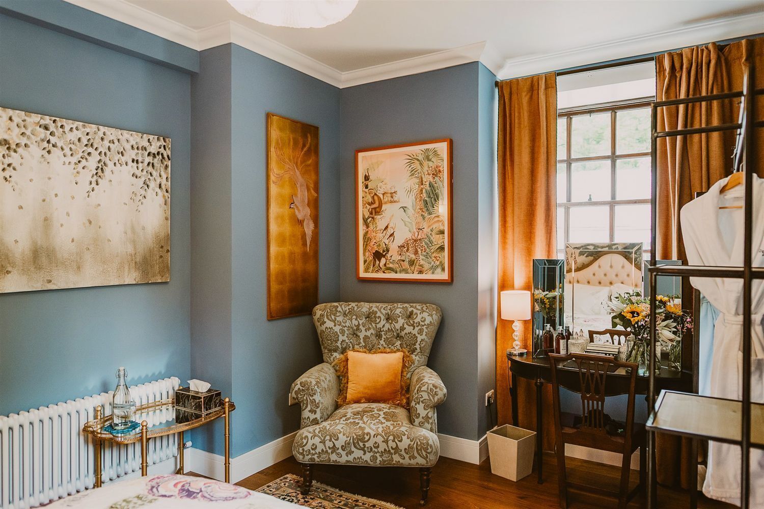 A room with a patterned armchair, a radiator, a table with a lamp, and a bed with a headboard.