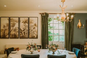 Ein Esszimmer mit Tisch für vier Personen, umgeben von Stühlen, einer Kristallleuchter und gerahmten Pflanzenbildern an der Wand.