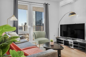A modern living room with a sofa, a green armchair, a TV, and a balcony with a view of buildings. - The Quay Waypost Hospitality Greenpoint (brooklyn)