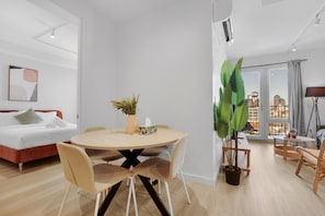 A modern living room with a round dining table, a sofa, and a large window offering a city view. - The Quay Waypost Hospitality Greenpoint (brooklyn)