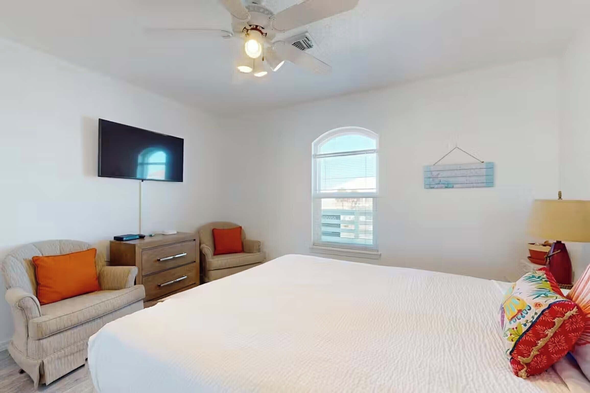 Une chambre à coucher comprenant un lit, une chaise, une commode, un téléviseur, un ventilateur de plafond, une fenêtre et un luminaire fixé au mur.