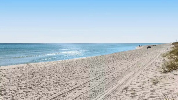 Une plage avec un ciel dégagé, un rivage sablonneux et des traces de pneus qui mènent à l’eau.