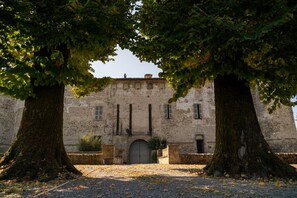 Un edificio histórico con una entrada central en arco, flanqueado por árboles grandes, y un camino de grava que lleva a la entrada.
