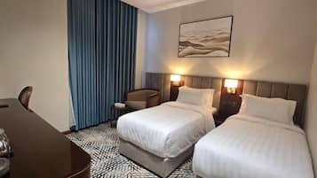A hotel room with two beds, a desk with a chair, a small table with a lamp, and a large window with curtains.