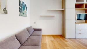 A modern living room with a grey sofa, wooden flooring, a TV on a wooden cabinet, and wall-mounted shelves. - Garda Hub Retreat by Booking Garda Lake (Puegnago sul Garda)
