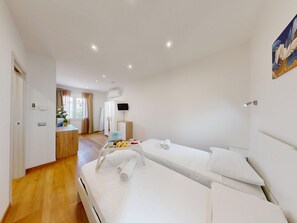 A hotel room with two beds, a desk with a flat-screen TV, a small table with fruits and cups, and a painting on the wall. - Garda Hub Retreat by Booking Garda Lake (Puegnago sul Garda)