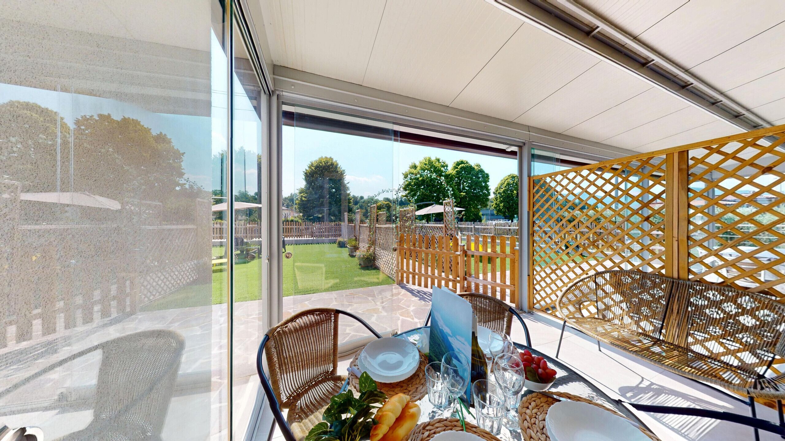 Un balcón con una mesa lista para comer, una silla de mimbre y vistas a un jardín con cerca de madera.