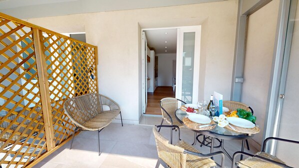 Un balcón con muebles de mimbre, una mesa de vidrio dispuesta para comer y vista al interior del edificio.