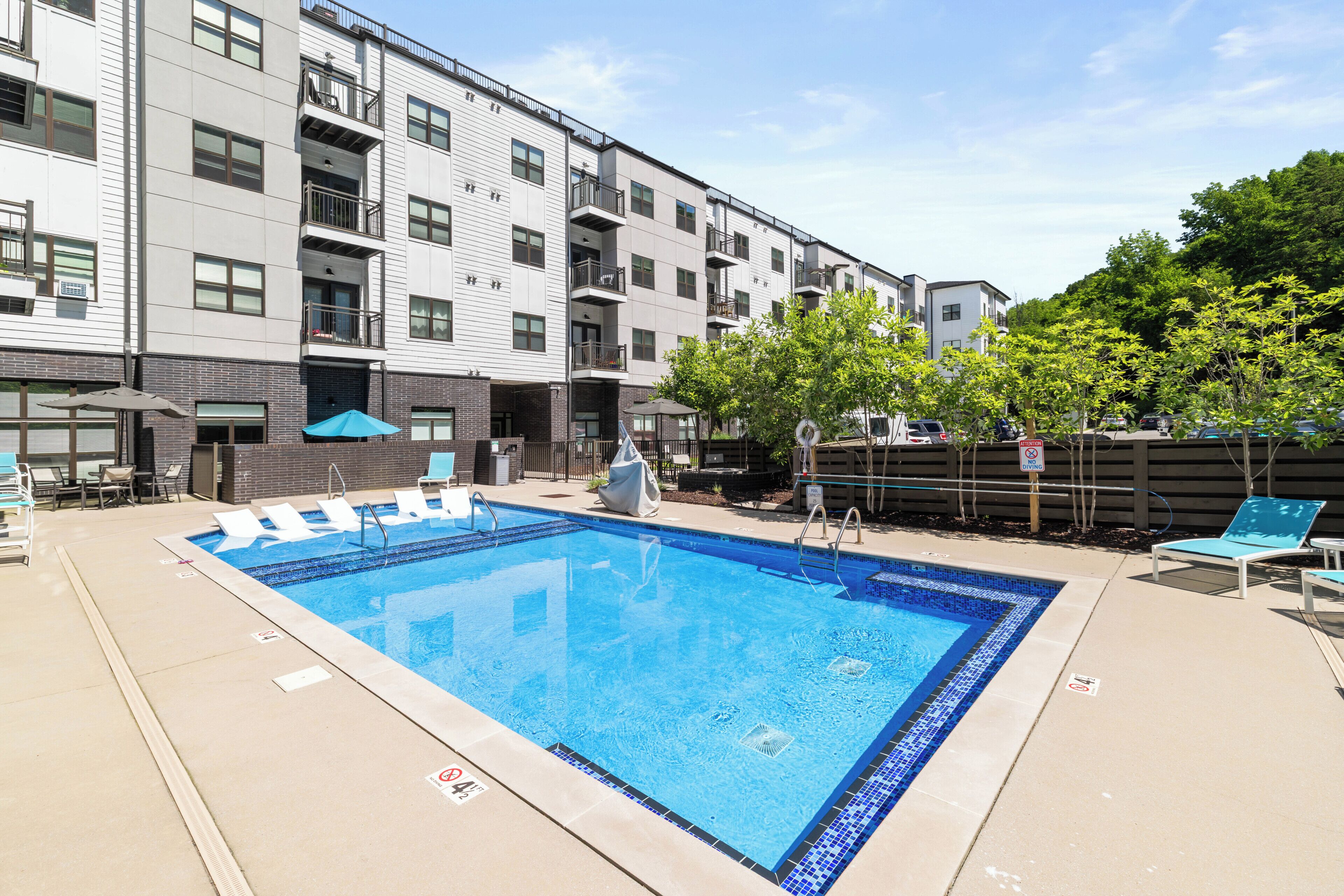 A rectangular pool with blue tiles, surrounded by lounge chairs and umbrellas, adjacent to a multi-story building with balconies.