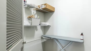 A laundry room with a metal rack, ironing board, shelves with folded clothes, and a fire extinguisher.