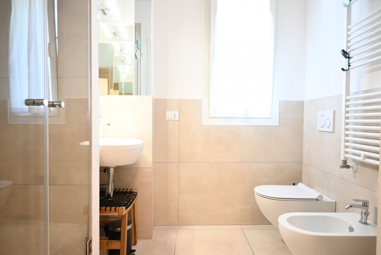 A modern bathroom with a glass shower enclosure, a sink, a bidet, and a window providing natural light.