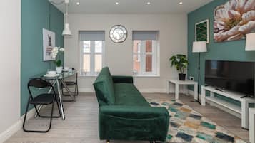 Ein modernes Wohnzimmer mit einem grünen Sofa, einem Essbereich mit Stühlen, einem Fernseher und einer Wanduhr.