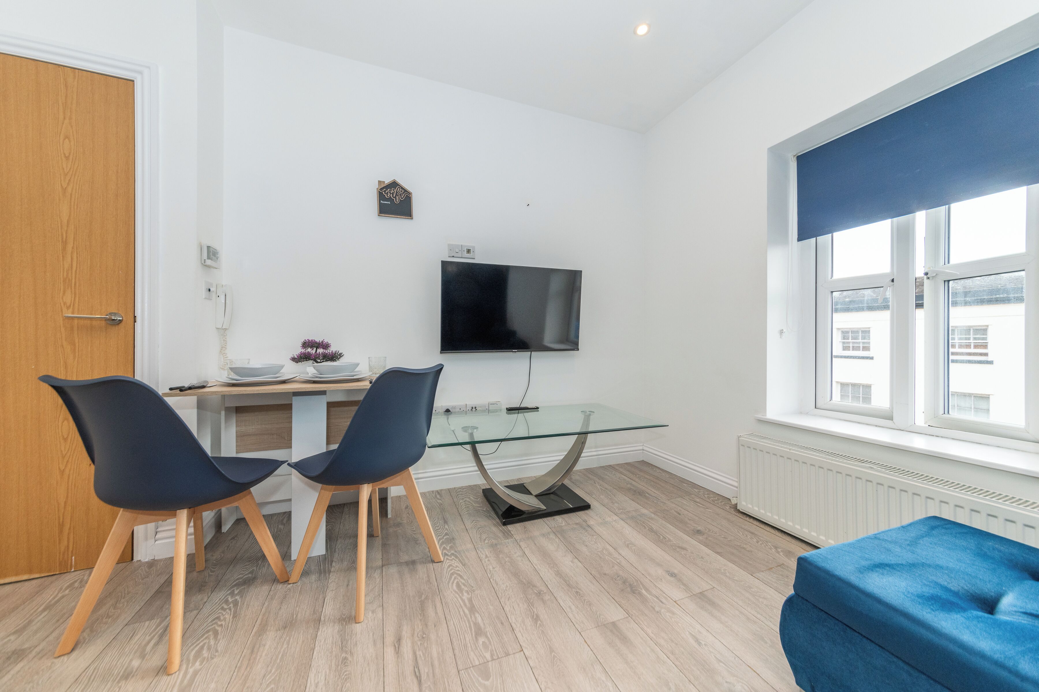 A modern living room with a glass dining table, two chairs, a blue sofa, a TV, and a window with blinds.