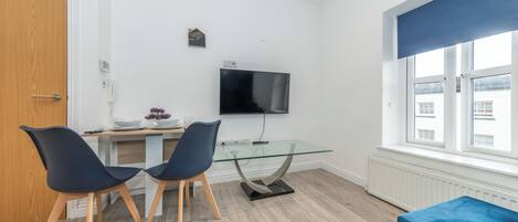 A modern living room with a glass dining table, two chairs, a blue sofa, a TV, and a window with blinds.