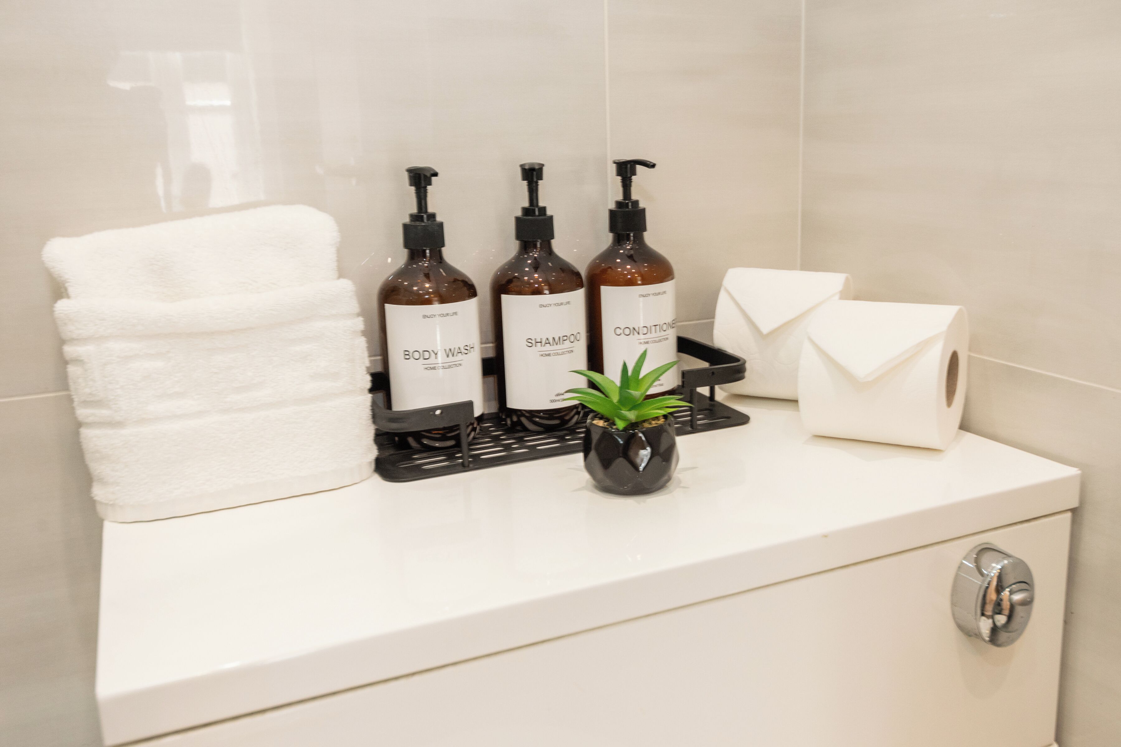 A bathroom with a white towel, three bottles of body wash, shampoo, and conditioner, a small potted plant, and a toilet paper holder.