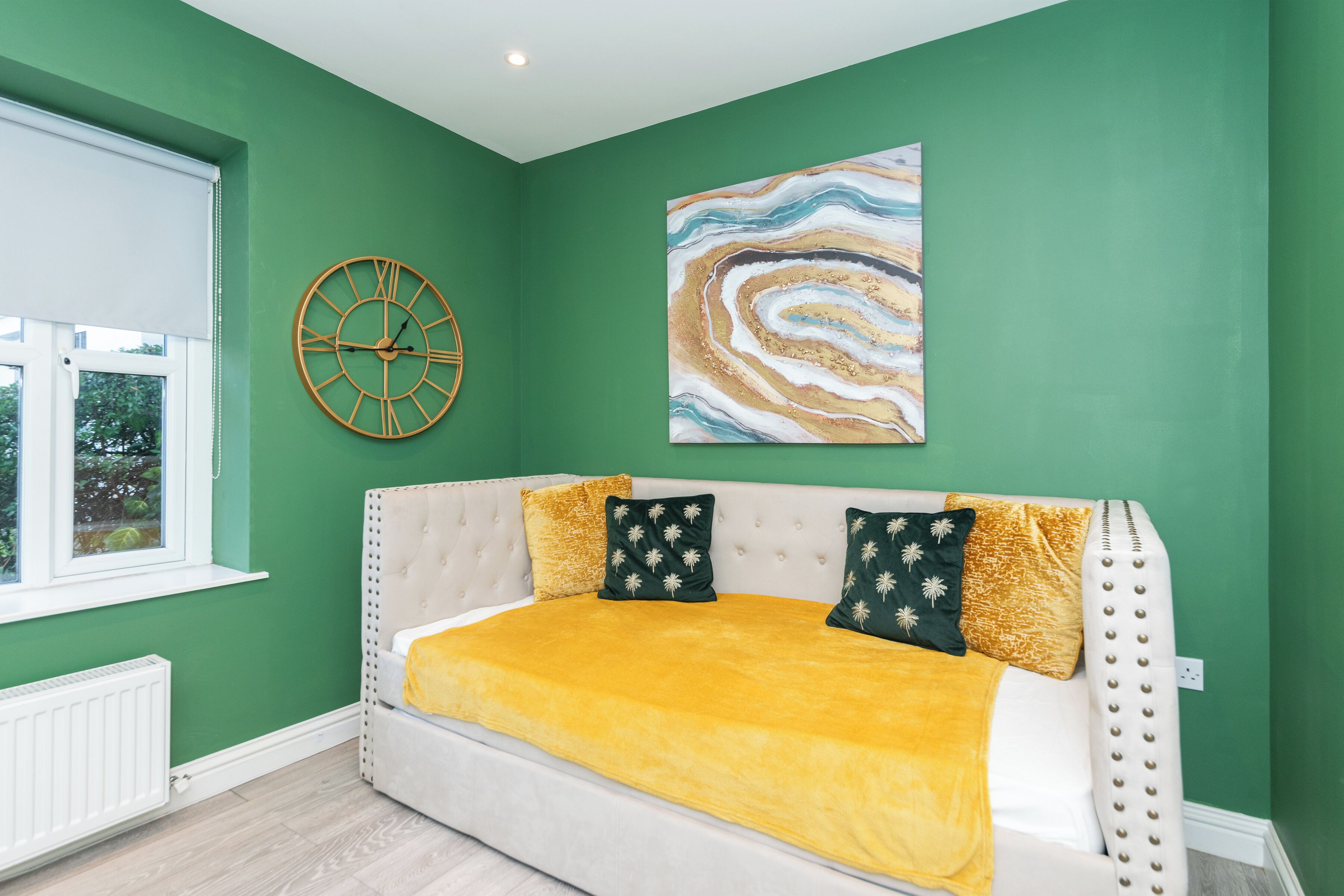 Una stanza con una parete verde, un divano bianco con una coperta gialla, una sveglia e un quadro con motivo marmorizzato.