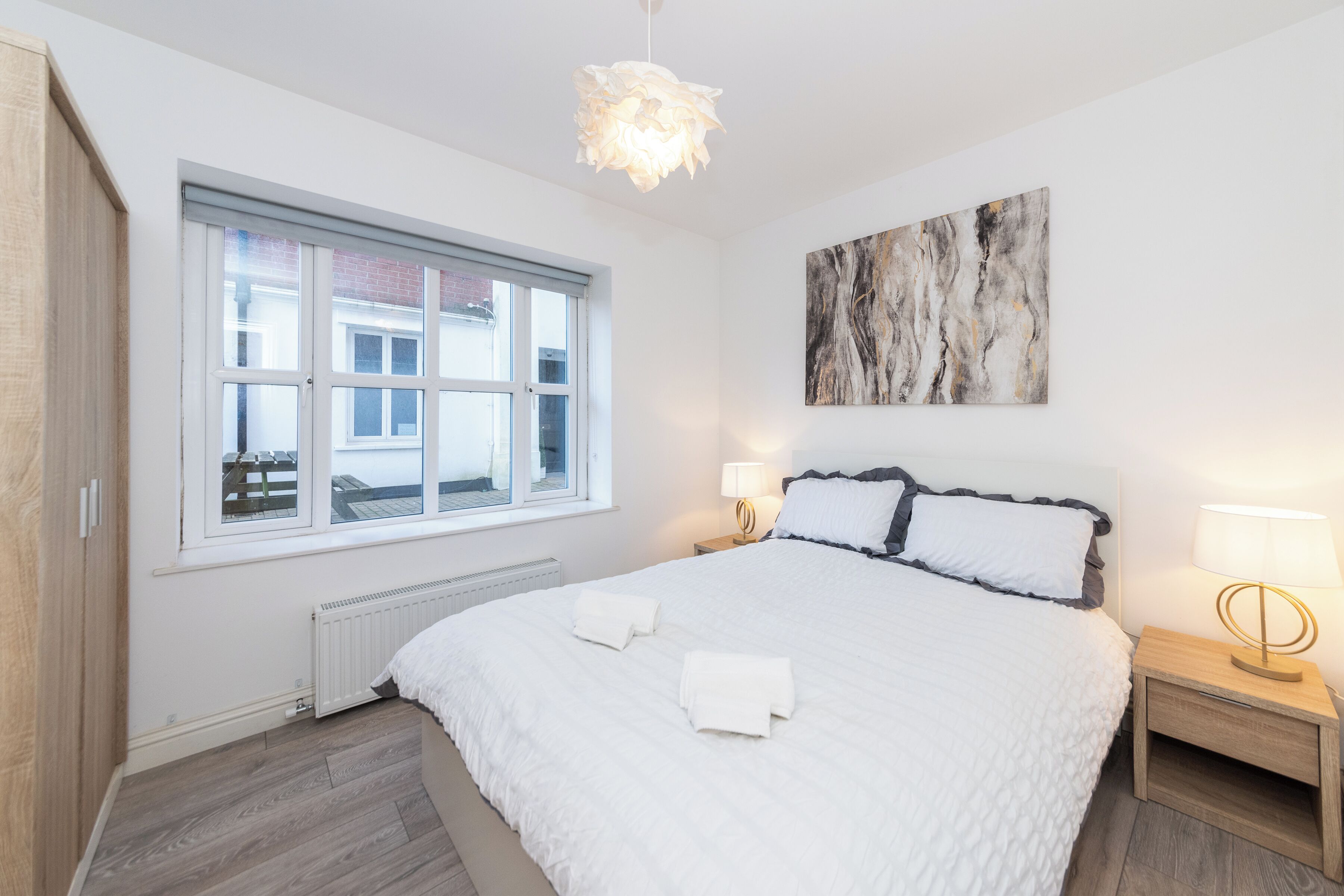 Un dormitorio con una cama grande, una ventana, un radiador, una mesita de noche con una lámpara y un cuadro en la pared.