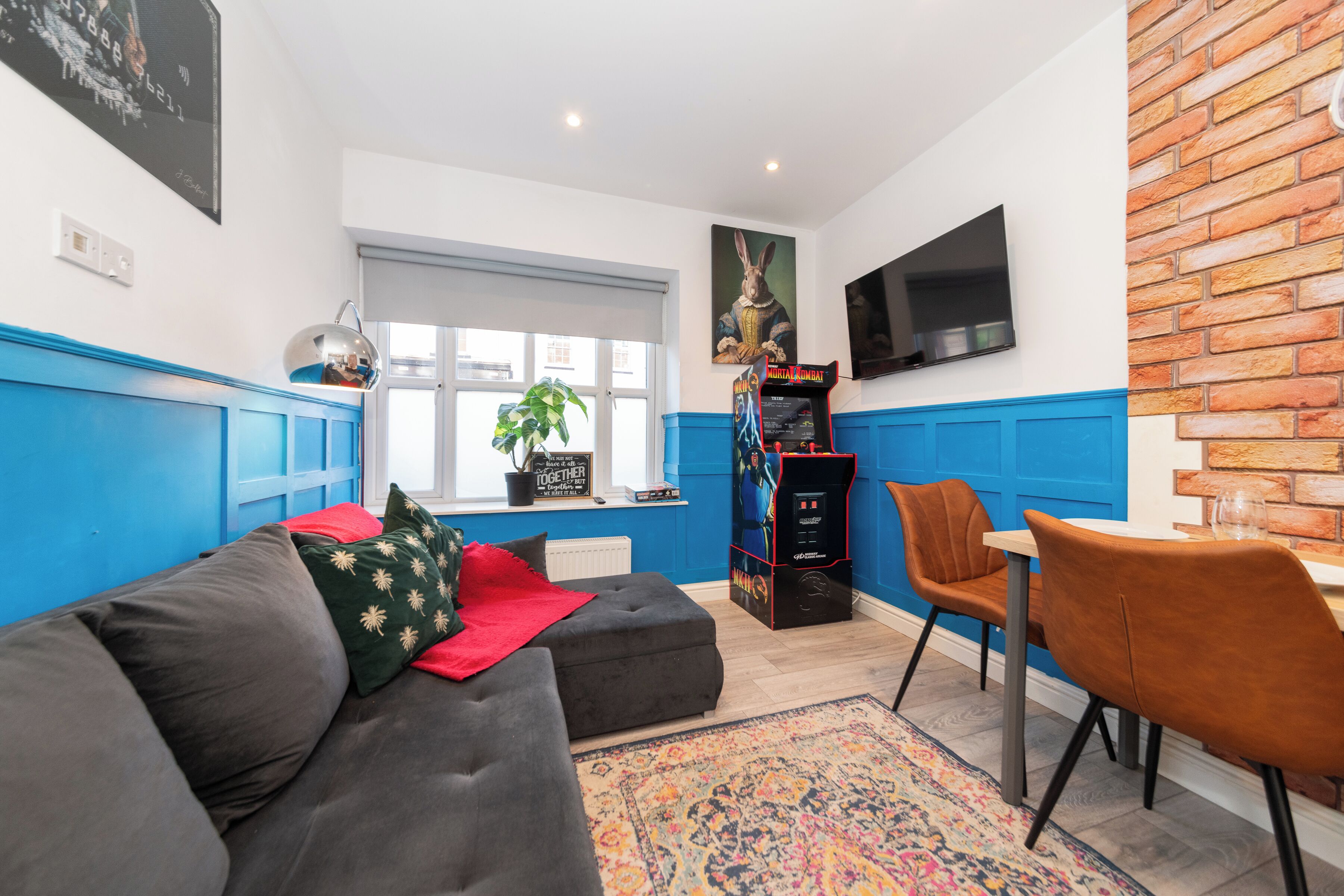 Ein Wohnzimmer mit Sofa, einem Esstisch mit Stühlen, einem an der Wand befestigten Fernseher und einer Zimmerpflanze.