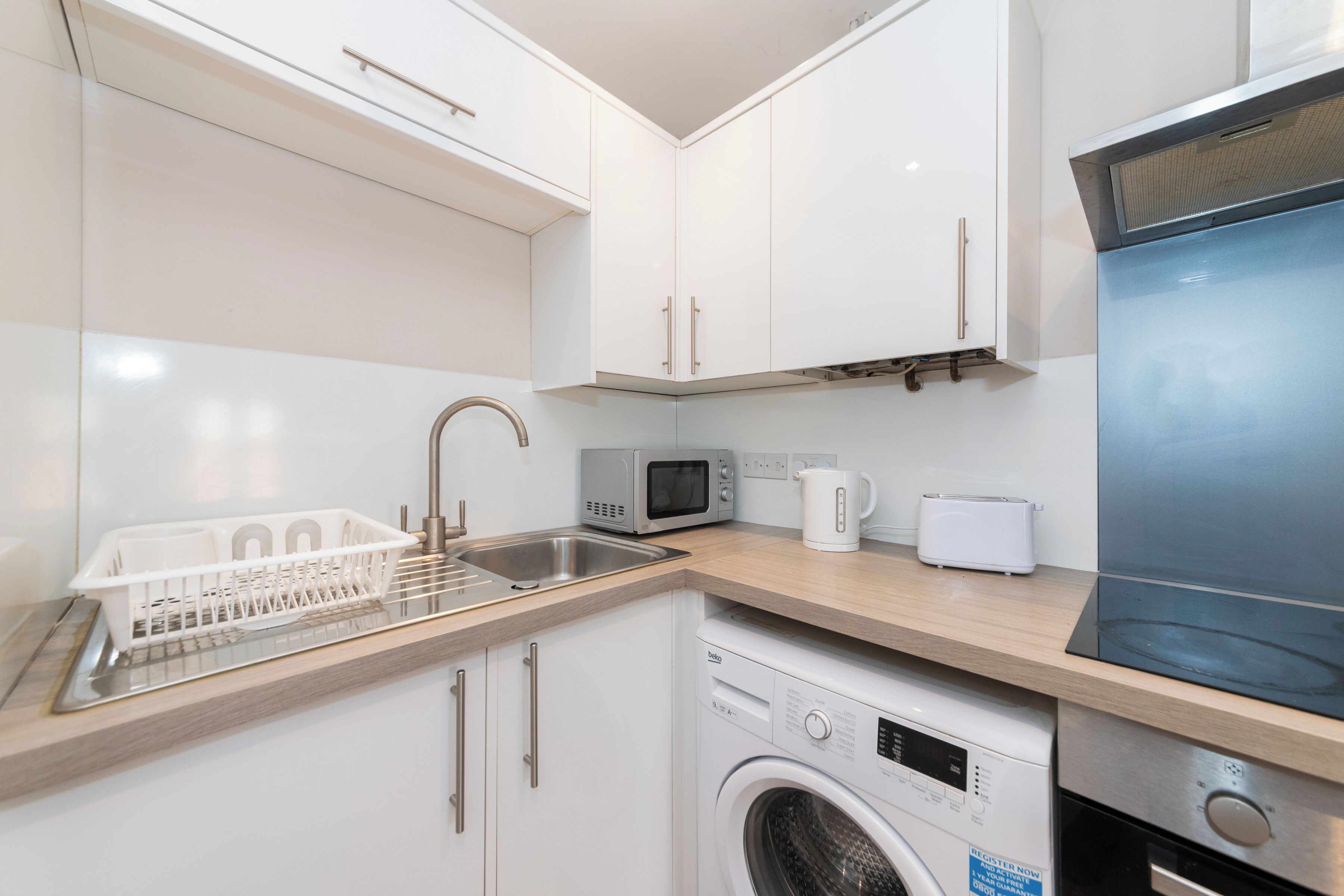 A compact kitchen with white cabinets, a stainless steel sink, a microwave, a kettle, a toaster, and a dishwasher.