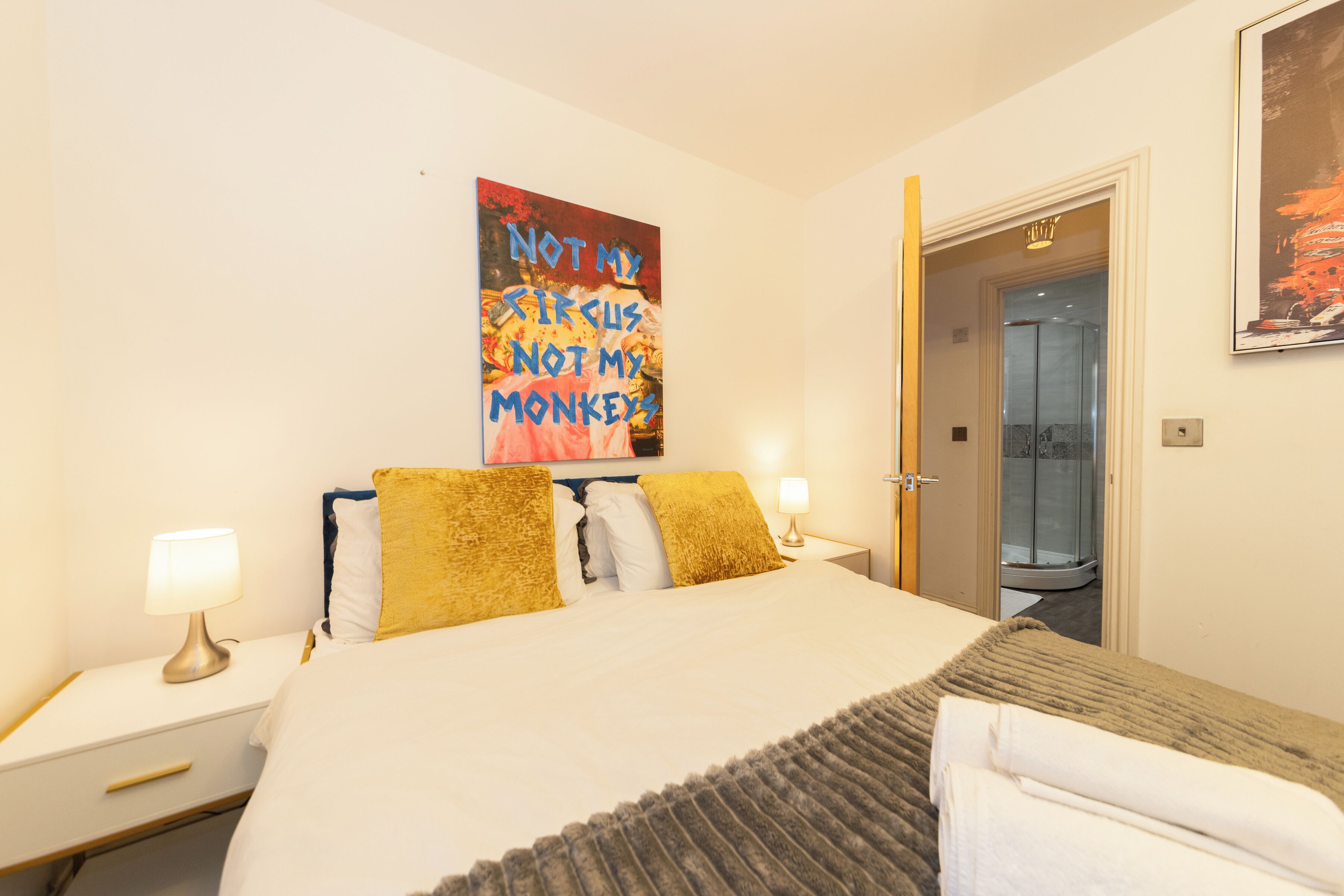 A hotel room with a bed, two bedside lamps, a poster on the wall, and a door leading to a shower area.