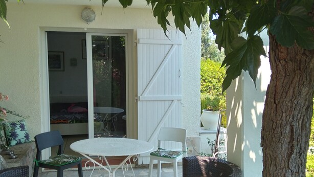 En udendørs terrasse med et bord og stole, et overdækket siddeområde og et træ der giver skygge.
