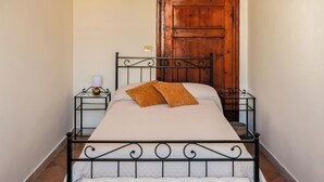 Uma cama com estrutura de metal, uma porta de madeira e uma pequena mesa de cabeceira com uma luminária.