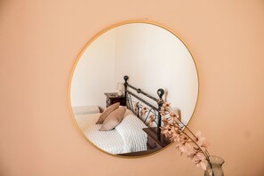 Un espejo circular en una pared color melocotón, reflejando una cama con cabecero blanco, una mesita de noche con una lámpara y un florero con flores.