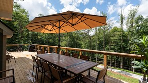 Eine Terrasse im Freien mit Holztisch und -stühlen, einem großen Sonnenschirm und Blick auf eine bewaldete Landschaft.