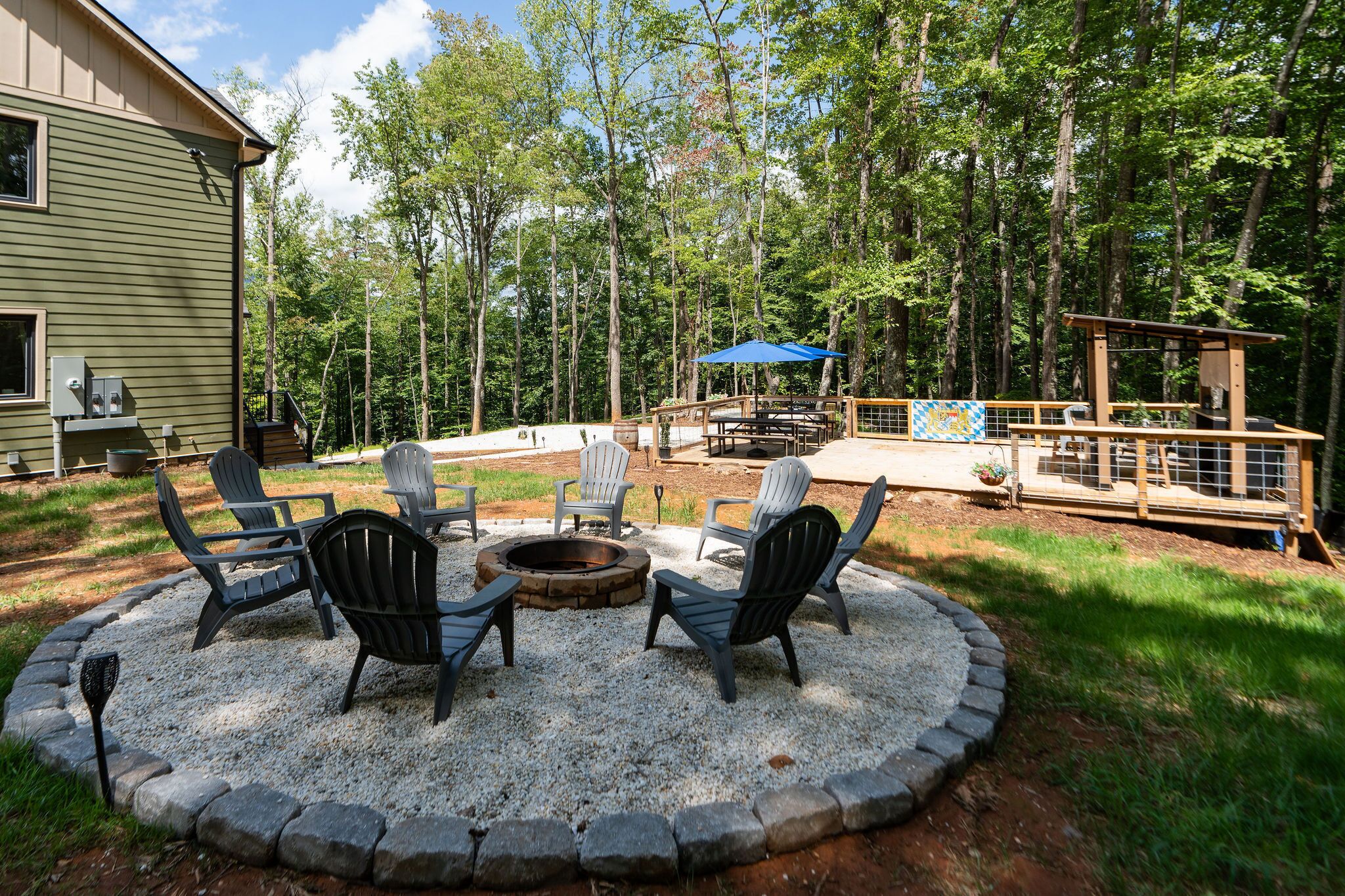 A circular seating area with Adirondack chairs around a fire pit, a deck with a blue umbrella, and a wooden structure in a wooded setting.