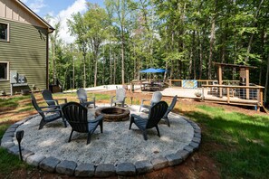 Un'area salotto circolare con poltrone Adirondack attorno a un braciere, un patio con un ombrellone blu e una struttura in legno immersa nel bosco.