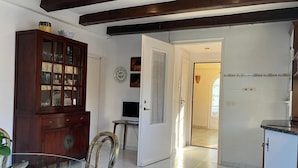 Uma área de refeições com mesa de vidro, cadeiras de madeira, um armário com portas de vidro e uma cozinha visível por uma porta aberta.