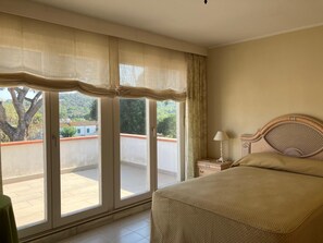 Ein Schlafzimmer mit Bett, Nachttisch, Lampe und Blick durch eine Schiebetür auf einen Außenbereich.