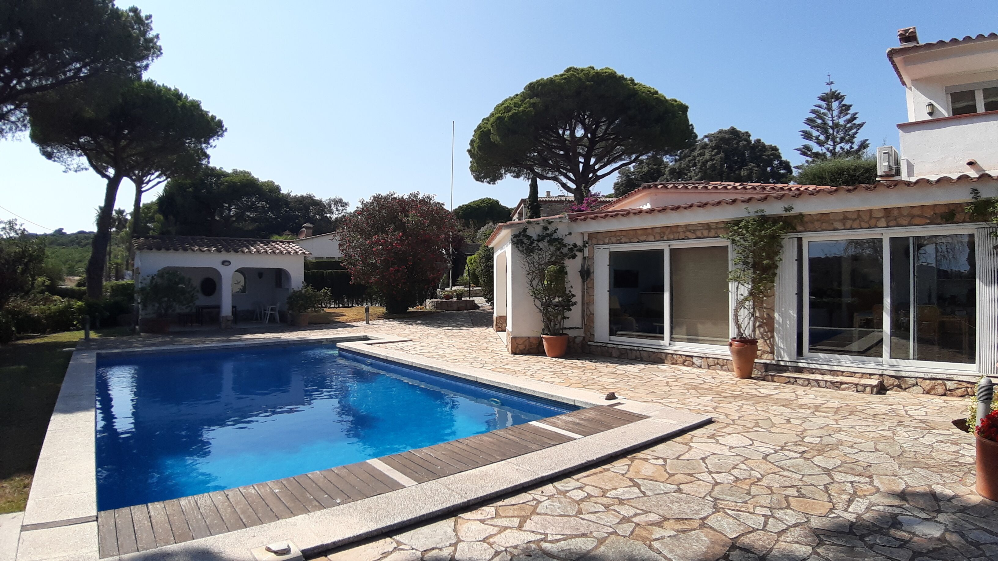 A swimming pool with a stone deck, a covered patio area, and a view of a residential property with a tiled roof and large windows.