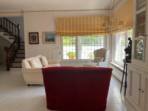 Ein Wohnzimmer mit Treppe, einem roten Sessel, einem weißen Sofa, einem Fenster mit Blick ins Grüne und einer Statue auf einem Sockel.