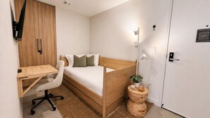 Una camera da letto con un letto di legno, una scrivania con una sedia, un tavolino rotondo con una pianta e una porta bianca con segnale di emergenza.