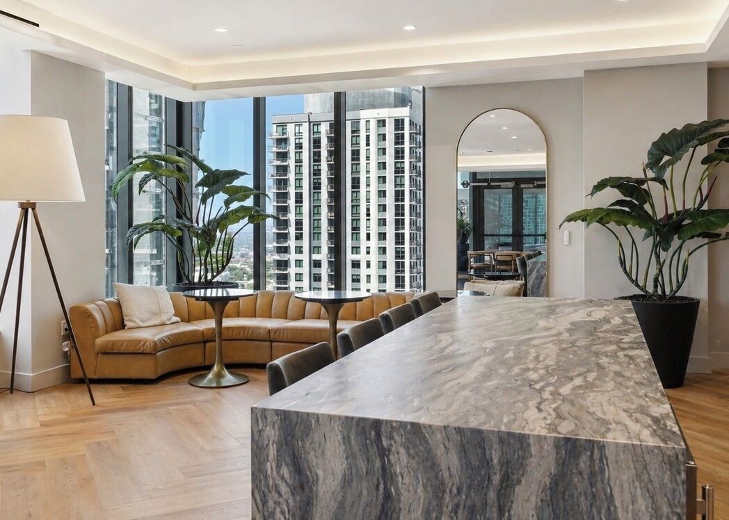 A modern living room with a marble dining table, a leather sofa, and large windows offering a city view.