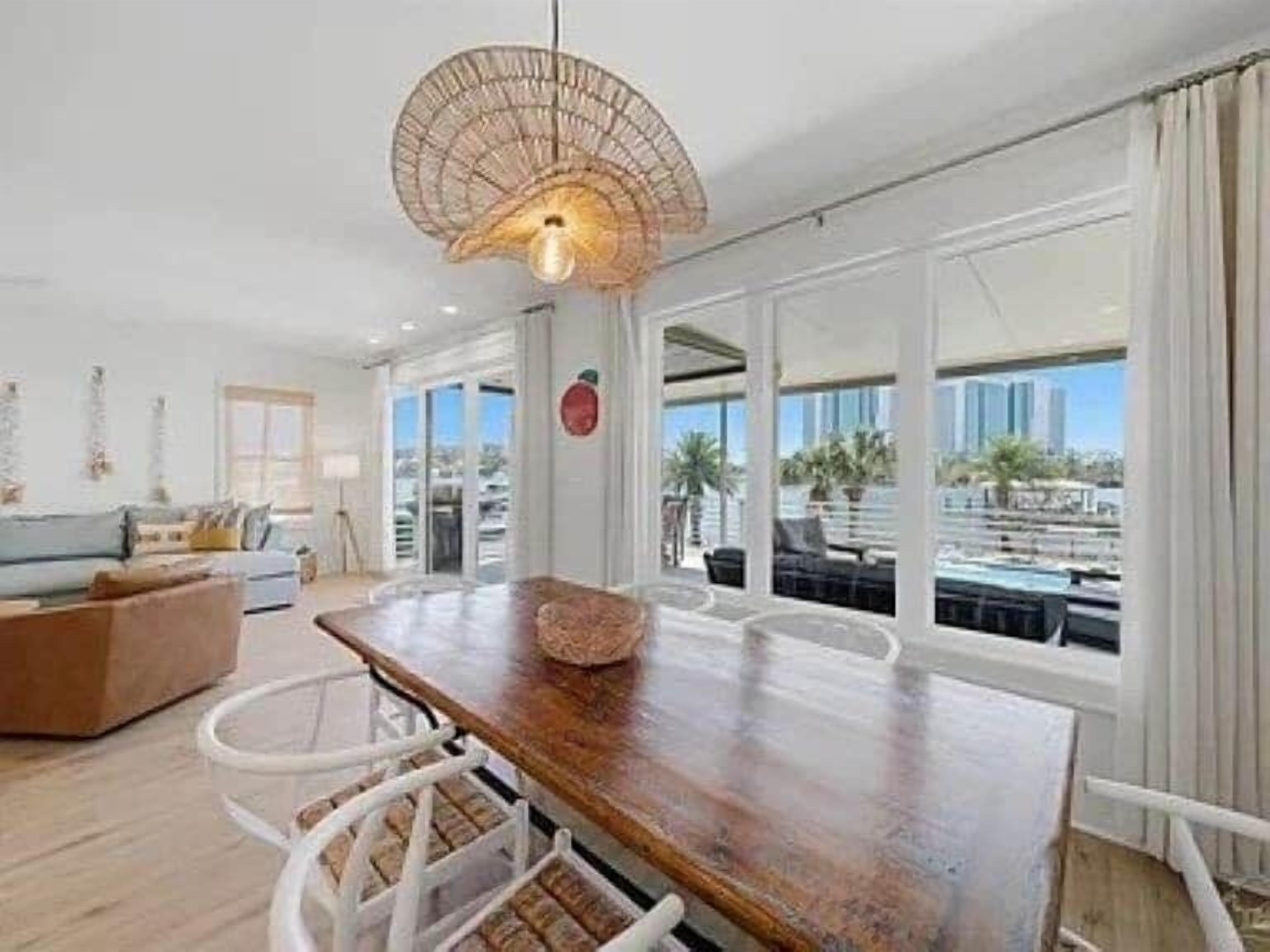 A spacious living area with a wooden dining table, white chairs, and a large window offering a view of palm trees and a cityscape.