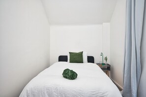A minimalist hotel room with a single bed, a nightstand, a green pillow, and a green plant. - Modern 2BR Mews House - Elizabeth Line | Sleeps 5 (Ilford)