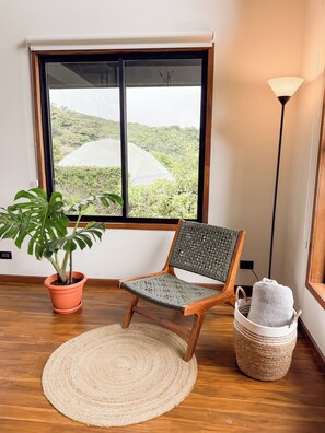A room with a wooden floor, a woven chair, a potted monstera plant, a round woven rug, a window with a view of green hills, and a floor lamp.